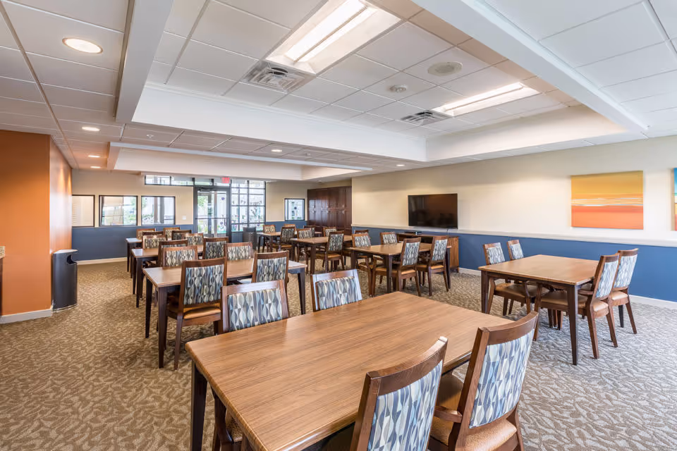 A spacious dining room with multiple wooden tables and patterned cushioned chairs arranged neatly. The room has a carpeted floor, a ceiling with recessed lighting, and a large flat-screen TV mounted on the wall. There are colorful paintings on the walls and large windows letting in natural light near the entrance.