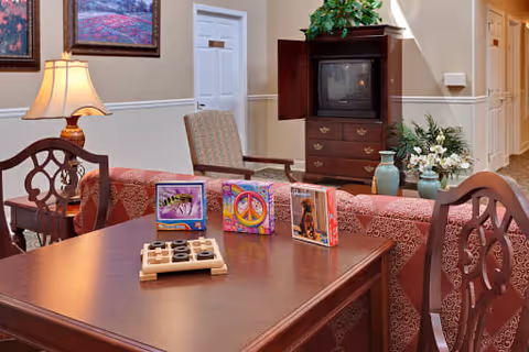 A cozy common area with a wooden table and chairs in the foreground, featuring a tic-tac-toe game and colorful decorative blocks. In the background, there is a patterned sofa, an armchair, a wooden cabinet with a TV, and a vase with flowers. The walls are beige with white trim, and framed artwork is hung on the wall.