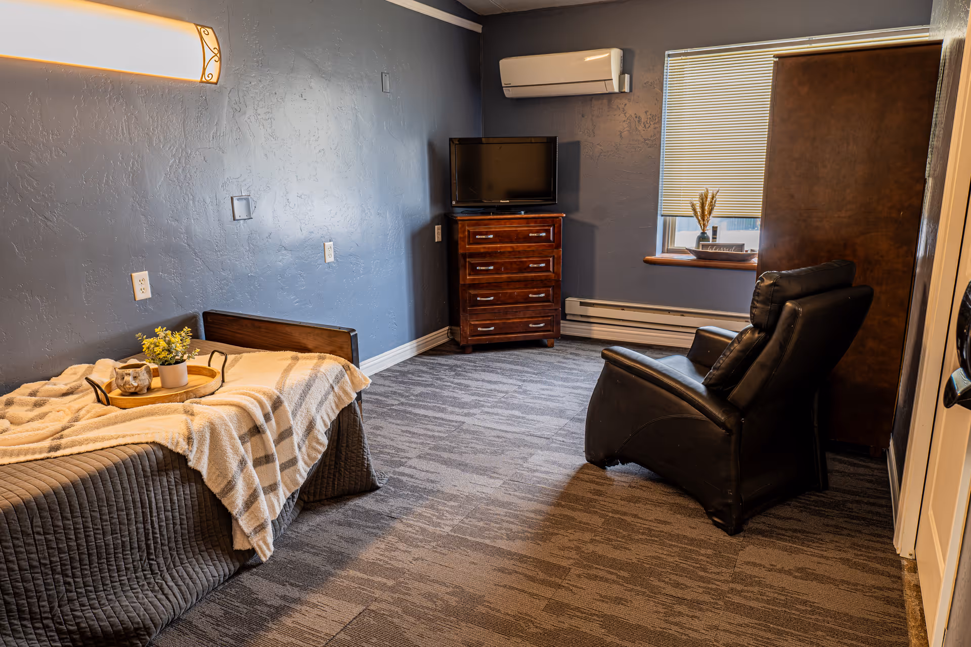 A cozy room with a single bed covered in a gray quilt and a beige striped blanket, a small tray with a potted plant and a decorative item on the bed. The room has blue textured walls, a dark wooden dresser with a TV on top, a black leather recliner chair, a window with blinds partially closed, and a wall-mounted air conditioning unit above the dresser.