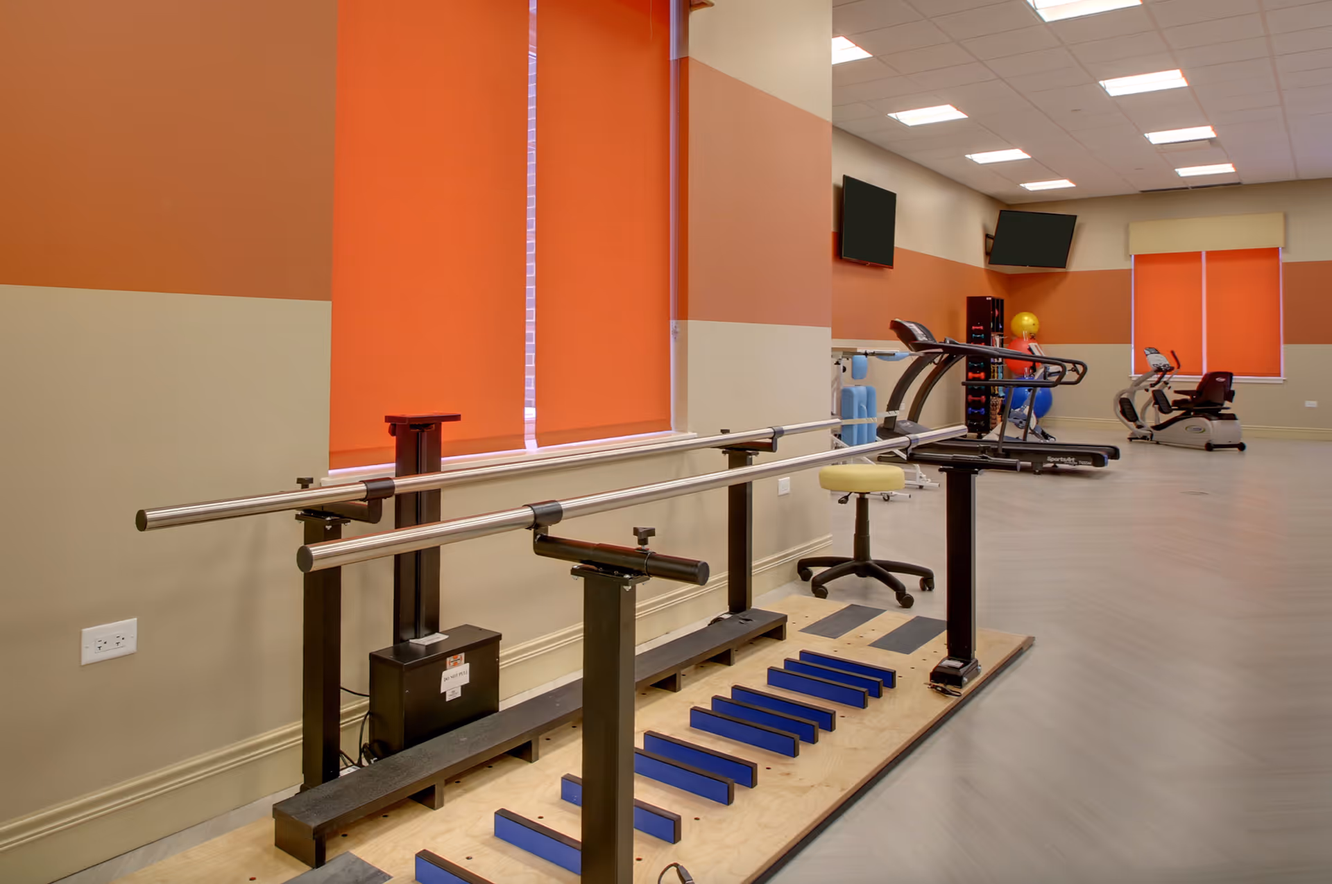 A rehabilitation or physical therapy room with parallel bars for walking exercises, a stool, treadmills, exercise balls, and stationary bikes. The walls are painted in two tones of beige and orange, with orange window blinds partially covering the windows.
