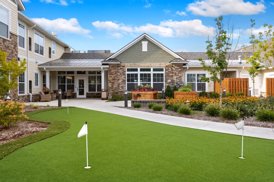 Outdoor courtyard area of a senior living facility with a putting green, landscaped garden beds, small trees, and a building with stone and siding exterior under a blue sky with some clouds.