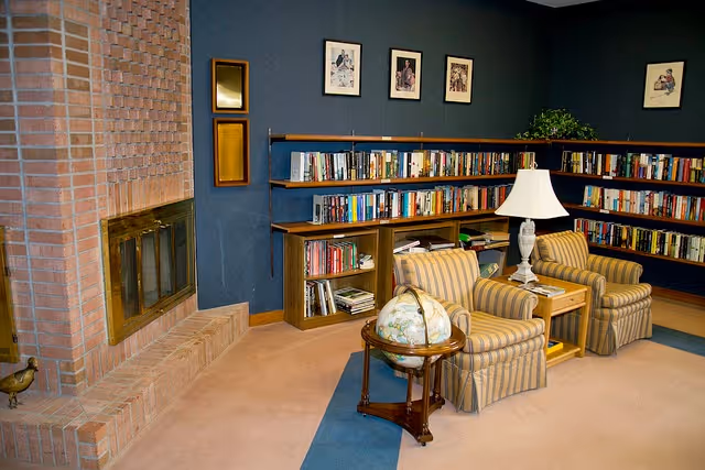 A cozy senior living facility library corner with a brick fireplace on the left, two striped armchairs facing each other with a small wooden table and a white lamp between them, a globe on a wooden stand in the foreground, and bookshelves filled with books along the dark blue walls. Framed pictures hang above the bookshelves.