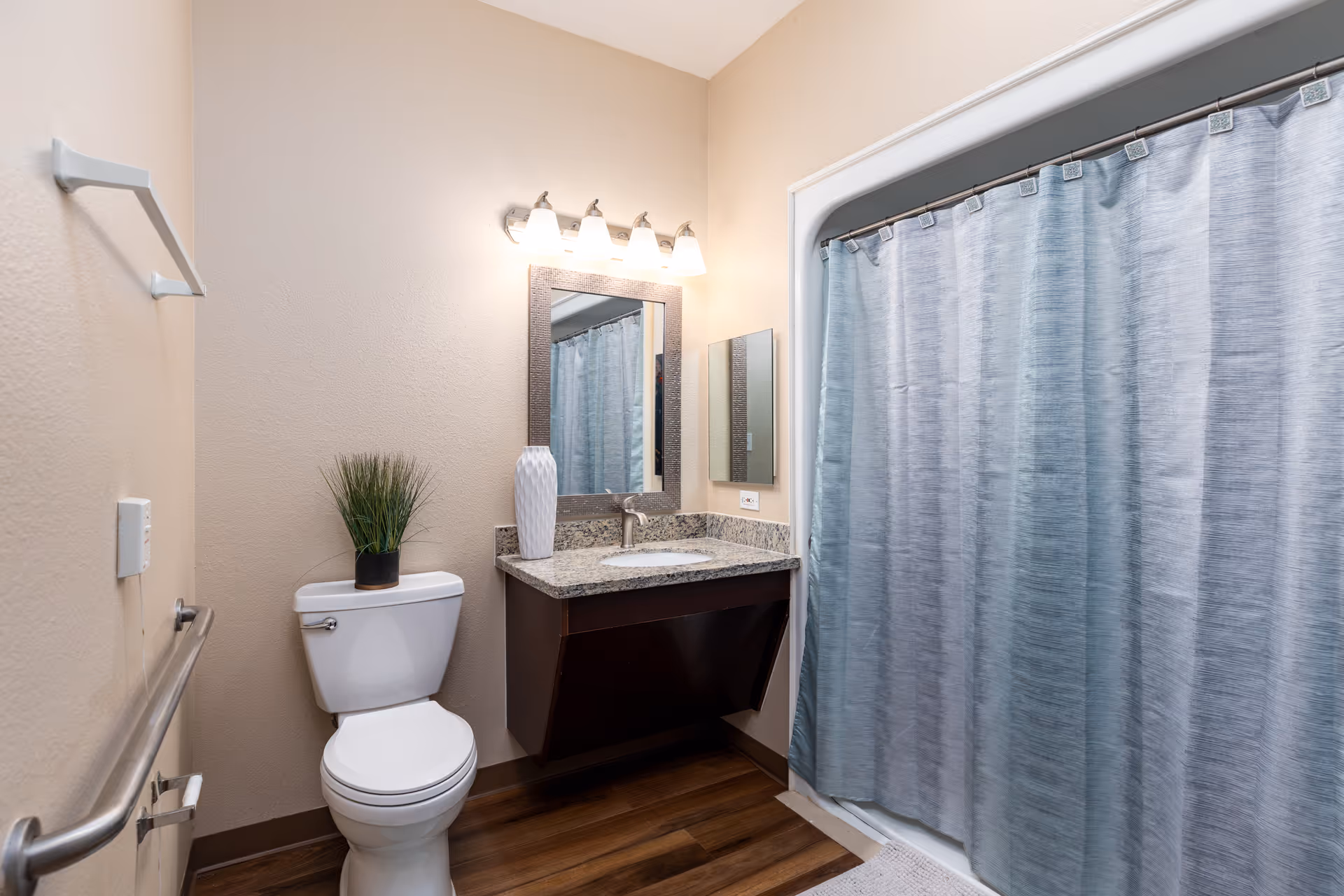 A clean and modern bathroom with a white toilet, a small potted plant on the toilet tank, a granite countertop with a sink, a large mirror with four light fixtures above it, and a shower with a blue curtain.