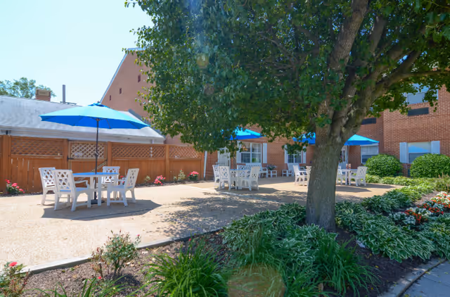 Outdoor patio area at Lakeside Senior Living with white tables and chairs under blue umbrellas, surrounded by a wooden fence, green bushes, flowers, and a large tree providing shade. The background shows a brick building under a clear blue sky.