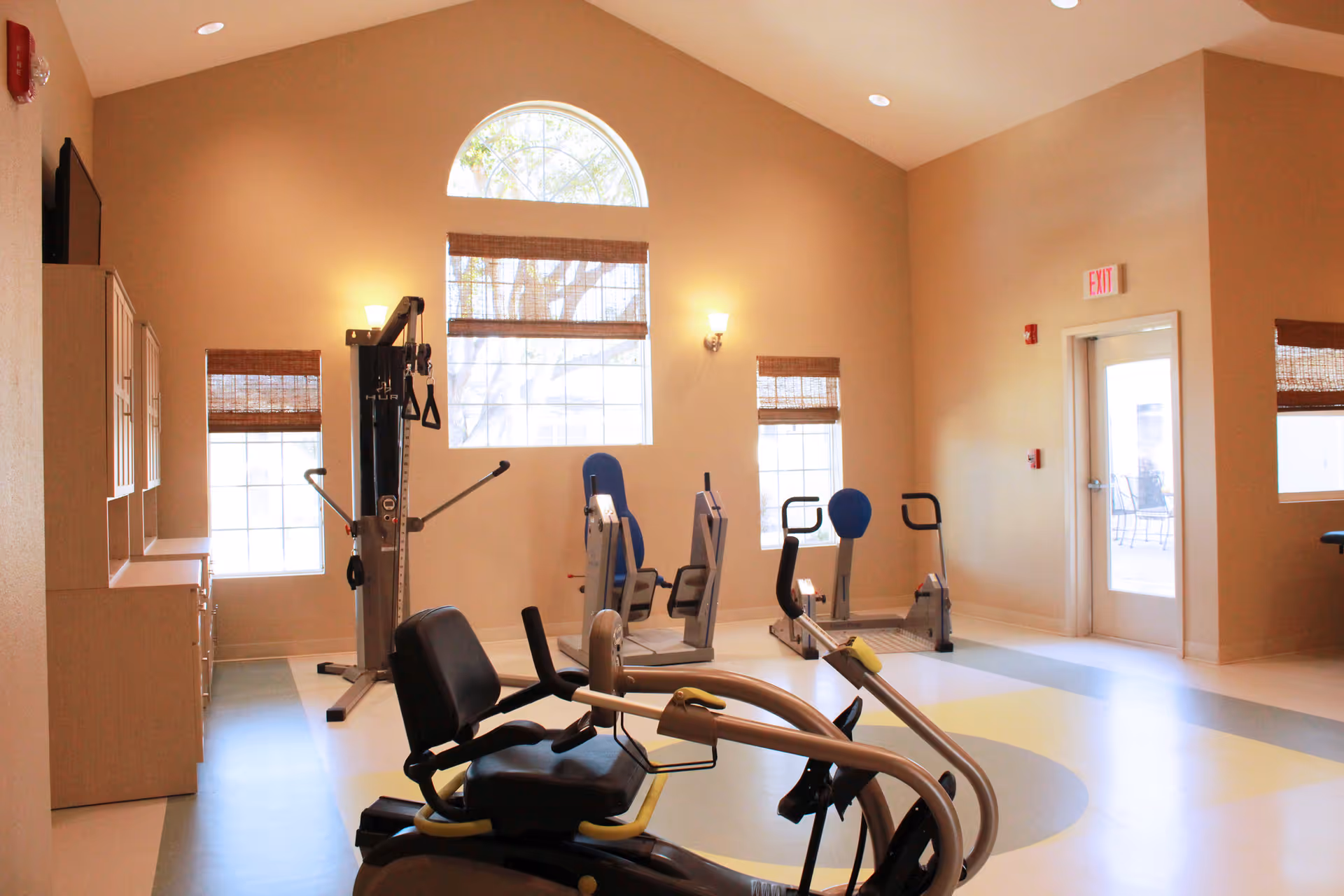 A bright rehabilitation exercise room with several pieces of fitness equipment including a recumbent bike, weight machine, and other exercise machines. The room has high ceilings, large windows with bamboo blinds, and light-colored walls and flooring. There is an exit door on the right side.