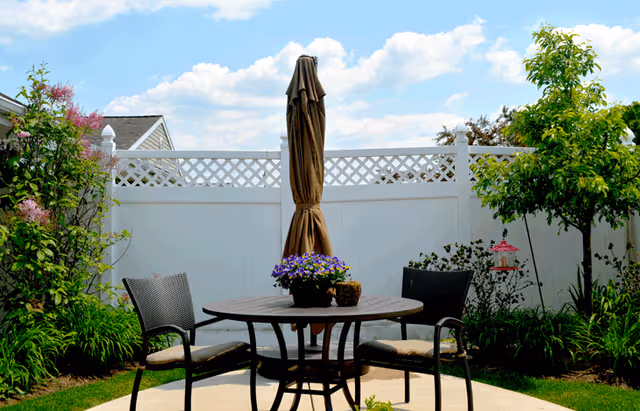 Outdoor patio area with a round table, two chairs with cushions, a closed umbrella in the center, surrounded by green plants and trees, and a white fence in the background under a partly cloudy sky.