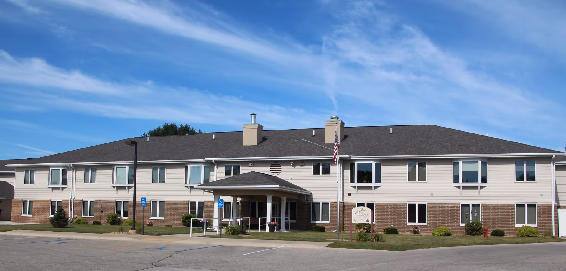 Front exterior of a two-story senior living community building with an entrance canopy, flagpole, and parking area.