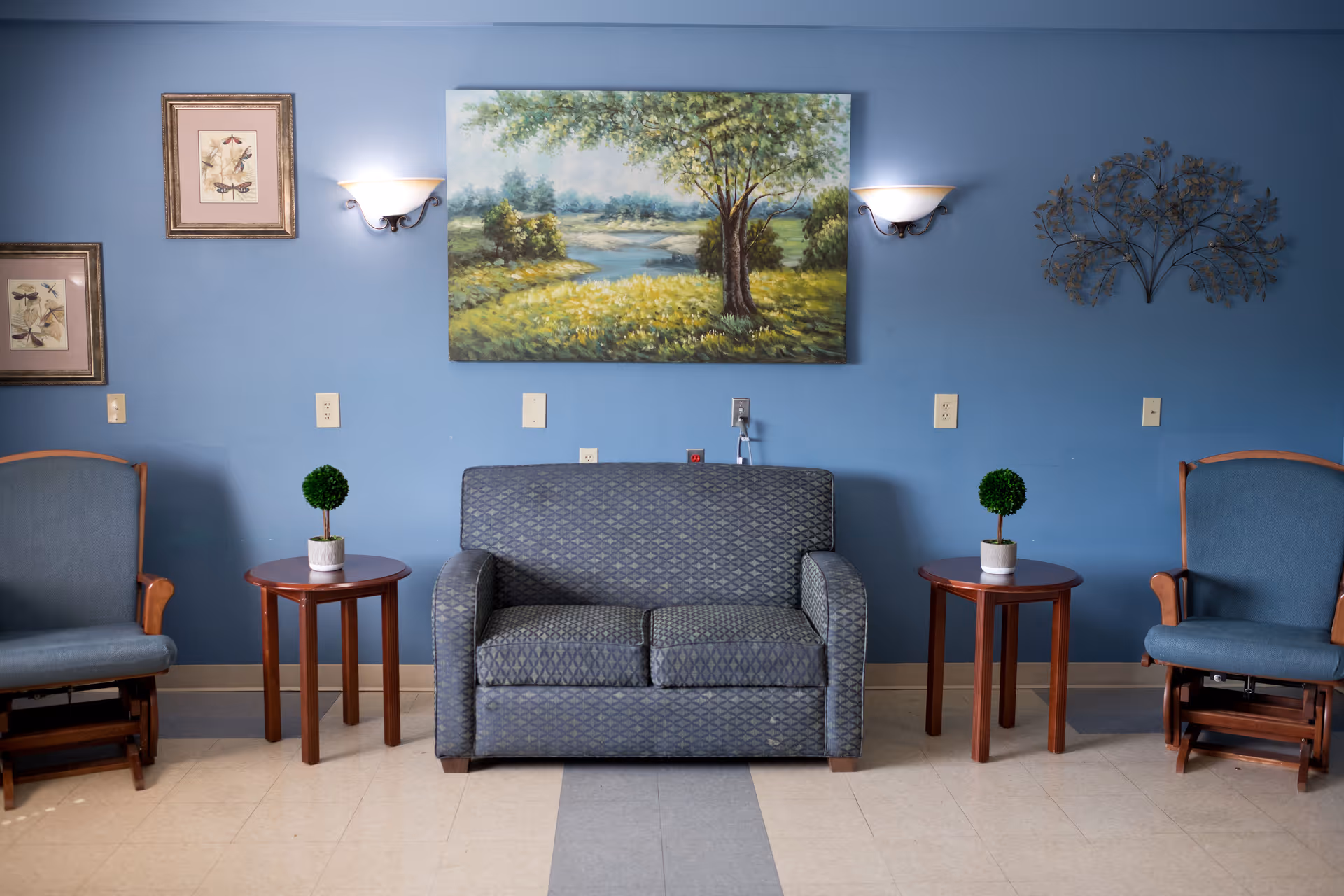 A sitting area with a blue patterned loveseat centered between two wooden side tables, each holding a small potted plant. On either side of the tables are two blue cushioned wooden armchairs. The wall behind is painted blue and decorated with a large landscape painting of a tree and greenery, two framed botanical prints, two wall sconces, and a metal wall art piece.