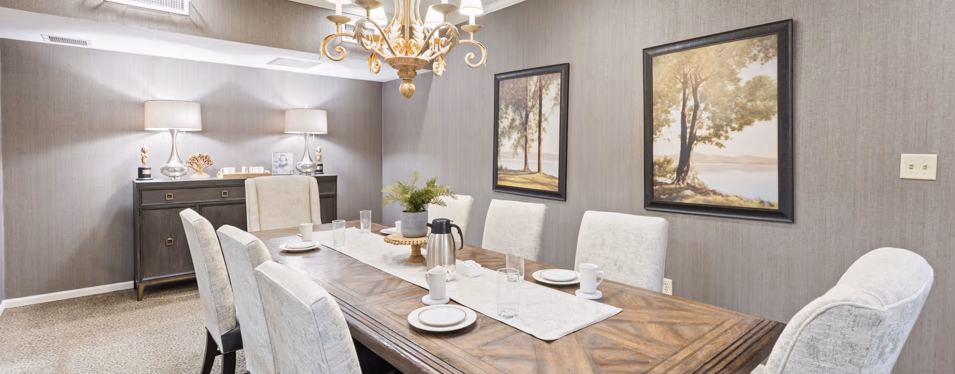 A dining room with a wooden table set for six people, featuring white plates, cups, and glasses. The table has a white runner and a small plant centerpiece. The room has beige upholstered chairs, two framed landscape paintings on the wall, a chandelier hanging from the ceiling, and a sideboard with two lamps and decorative items.