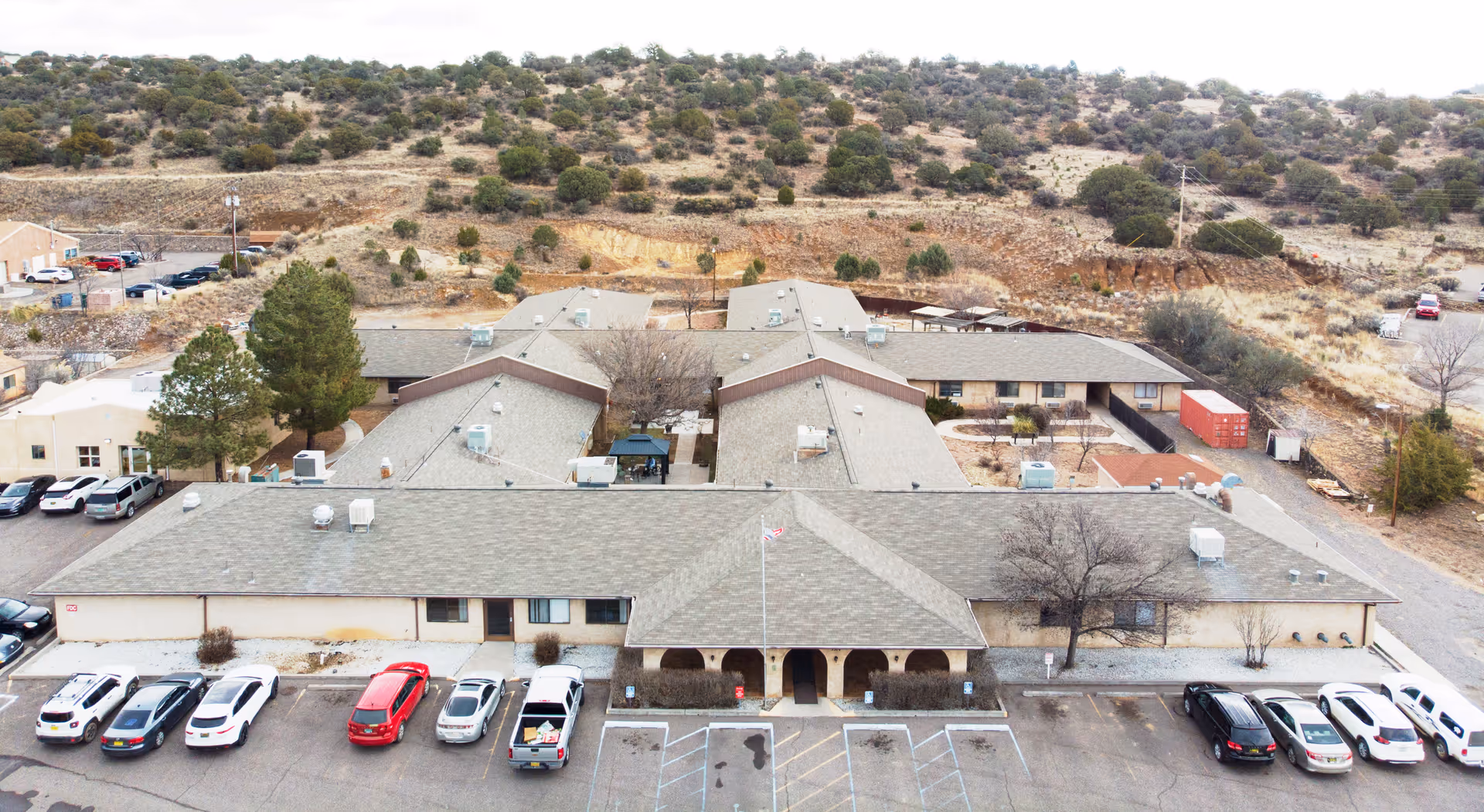 Aerial view of a single-story senior living facility building with a parking lot in front, surrounded by a dry, shrub-covered landscape with hills in the background. Several cars are parked in the lot, and the building has a central entrance with a covered porch and a flagpole.