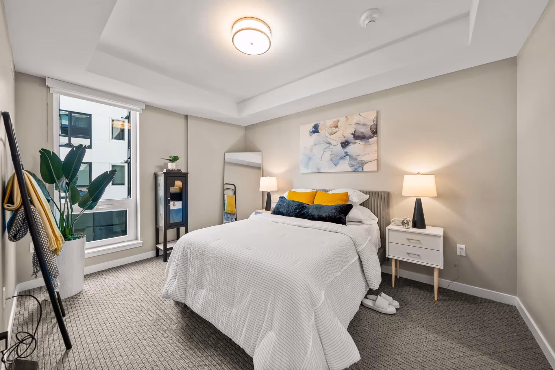 A neatly arranged bedroom with a bed covered in white bedding and accented with yellow and dark blue pillows. There are two bedside tables with lamps, a large window with a view of an adjacent building, a tall mirror leaning against the wall, a small cabinet with a plant on top, and a coat rack with hanging scarves. The room has beige walls and carpeted flooring.