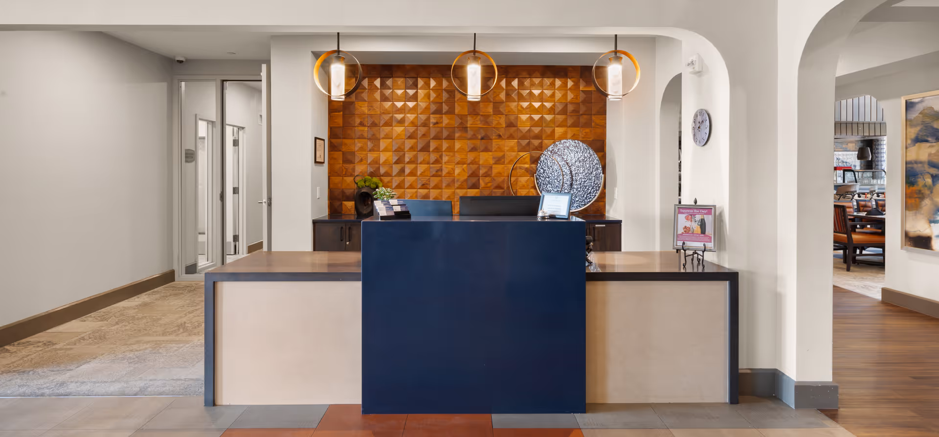 Reception desk area in a senior living facility with a dark blue front panel and beige sides. Behind the desk is a wooden textured wall with geometric patterns and three hanging pendant lights. To the right, there is a hallway leading to a dining area with tables and chairs. The floor is tiled with a mix of colors and materials.