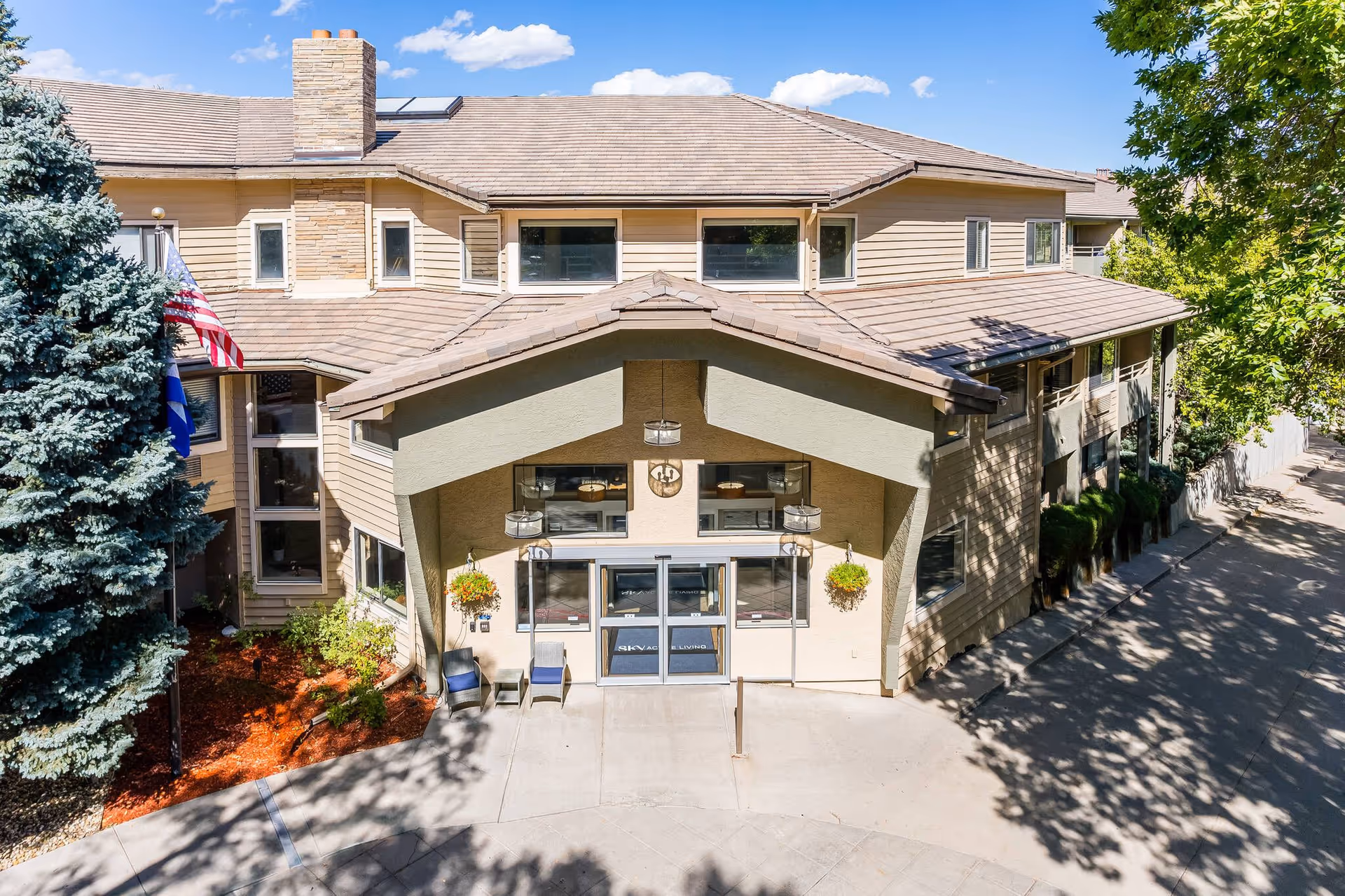 Front exterior view of a two-story senior living facility building with beige siding and a peaked roof. The entrance has double glass doors with a covered porch area, two chairs, hanging flower baskets, and an American flag on the left side. Trees and landscaping surround the building under a clear blue sky.