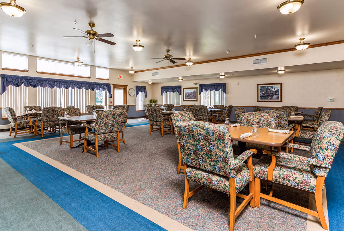 A spacious dining room with multiple wooden tables and floral upholstered chairs arranged neatly. The room has large windows with vertical blinds and blue valances, ceiling fans, and framed artwork on the walls. The carpet has a patterned design with blue borders.