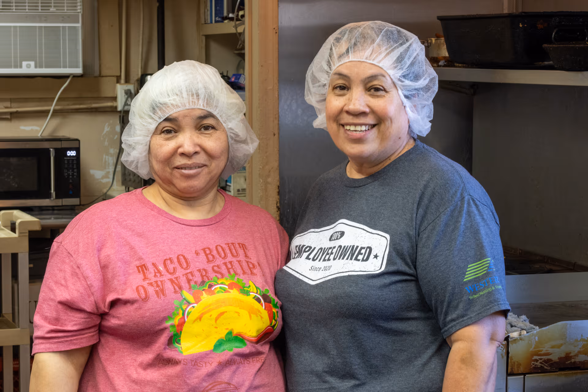 Two smiling women wearing hairnets pose in a commercial kitchen.