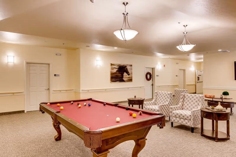A well-lit recreational room with a red felt pool table in the center, surrounded by beige walls and carpeted floor. There are two patterned armchairs and several wooden side tables with decorative items. The room features ceiling lights and a painting of a horse on the wall.