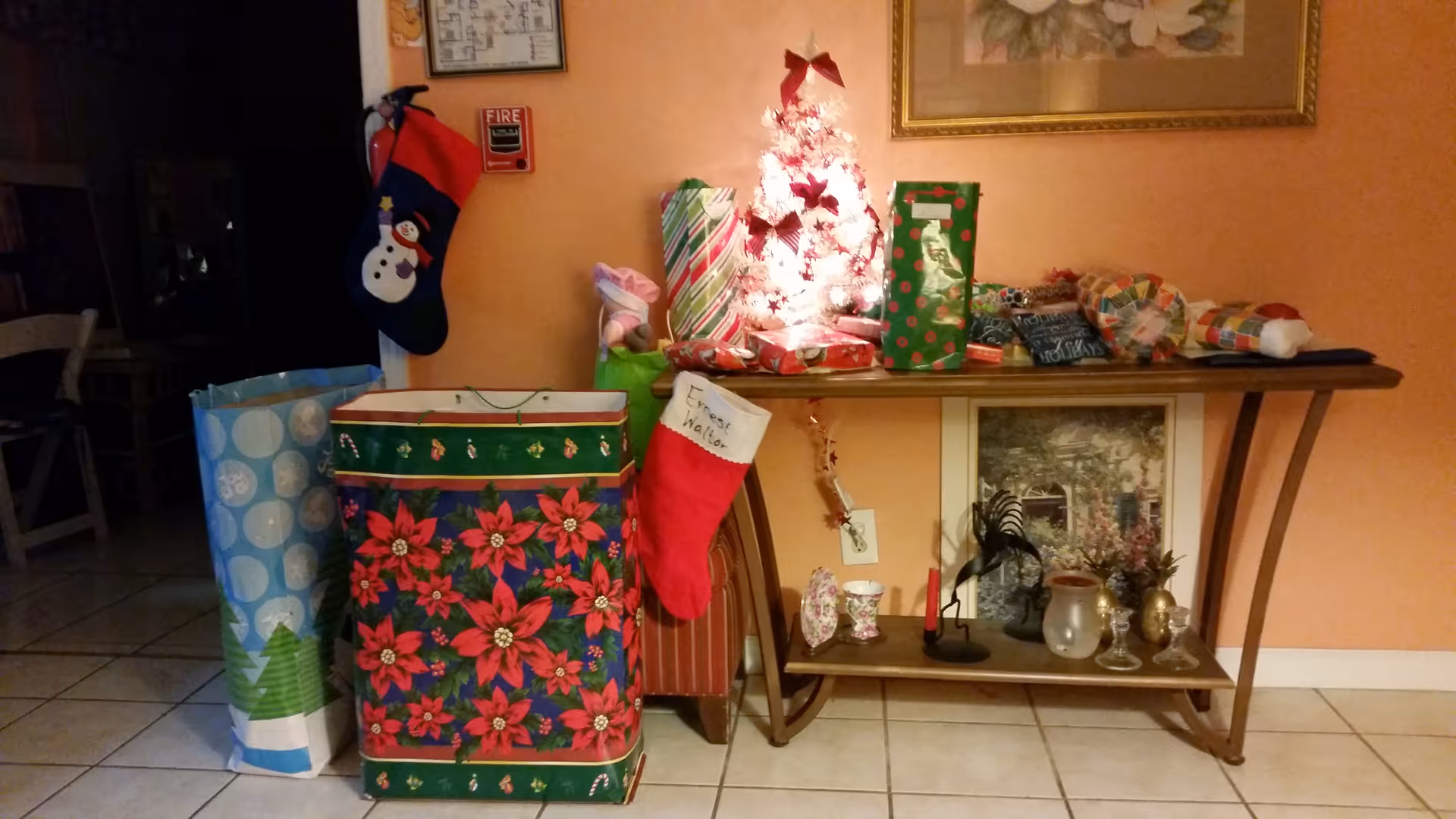 A festive Christmas display with wrapped gifts, a small lit tree, and a red stocking on a console table in a peach-colored living room.