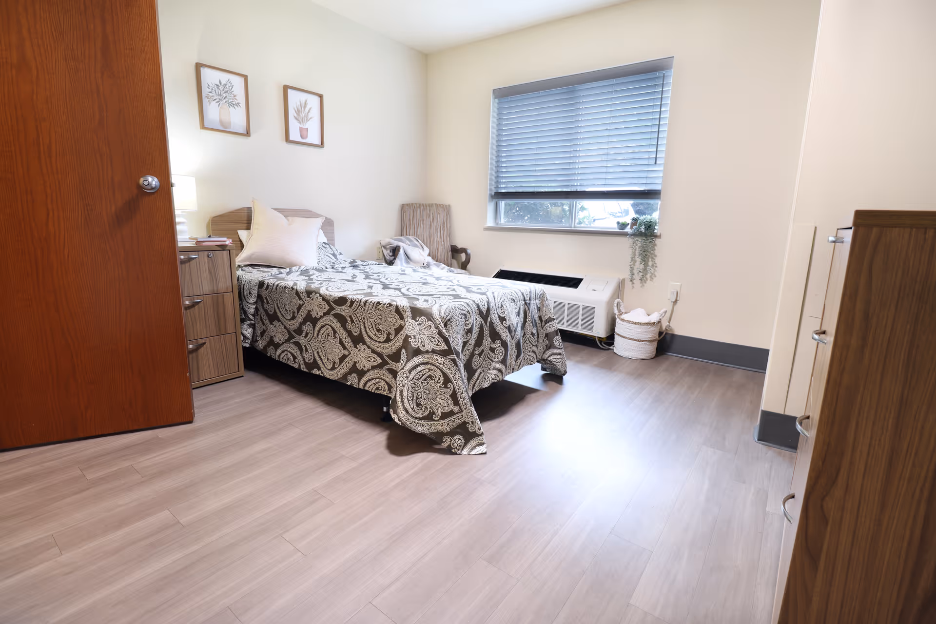 A simple, clean bedroom in a senior living facility with a single bed covered in a patterned bedspread, a wooden nightstand with a lamp, a wooden dresser, a chair near the window with blinds partially closed, and light-colored walls with two framed botanical prints.