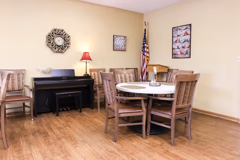 A cozy room with a round table surrounded by six wooden chairs with patterned cushions. Against the wall is a black upright piano with a small bench. A decorative wall clock, a red table lamp, and two framed pictures hang on the beige walls. An American flag stands in the corner near a wooden podium. The floor is wood laminate.