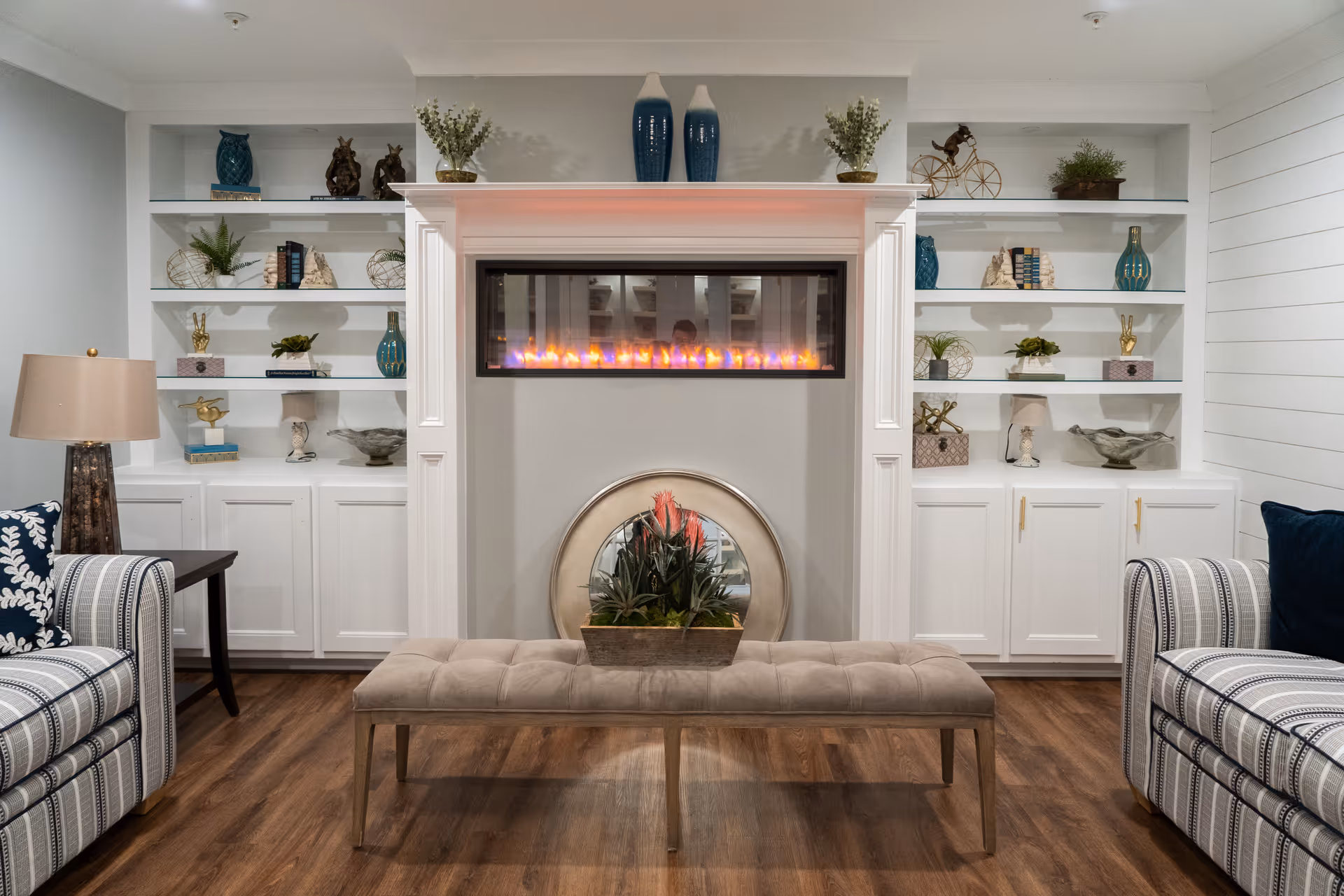 A cozy living room area with a modern electric fireplace centered between two white built-in shelves filled with decorative items. In front of the fireplace is a cushioned bench with a plant centerpiece. On either side of the bench are striped upholstered armchairs with patterned pillows. The room has wooden flooring and a table lamp on the left side.