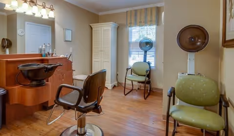 Interior view of a hair salon area in a senior living facility with a salon chair, a wooden cabinet with hair products, a white wardrobe, two green cushioned chairs, a window with striped curtains, and a vintage hair dryer.
