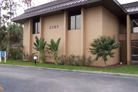 Beige River Commons building exterior with numbered facade, shrubs, and a paved driveway.
