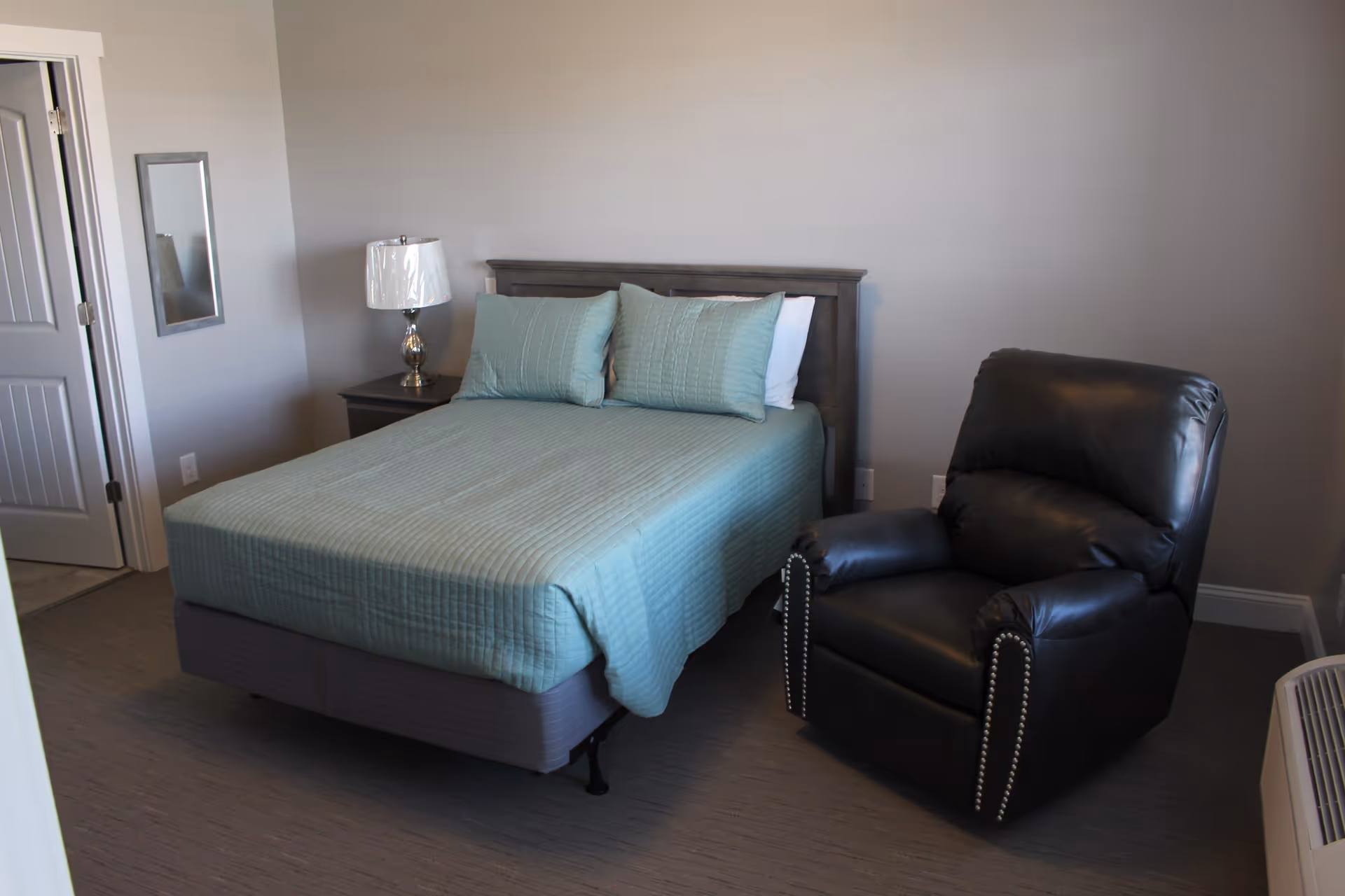 A bedroom with a neatly made bed covered with a light blue quilt and matching pillows, a wooden nightstand with a lamp, a black leather recliner chair, a wall-mounted mirror, and a partially open door.