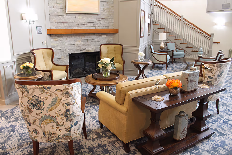 Cozy senior living lounge with upholstered chairs, sofas and side tables arranged around a fireplace and a staircase in the background.