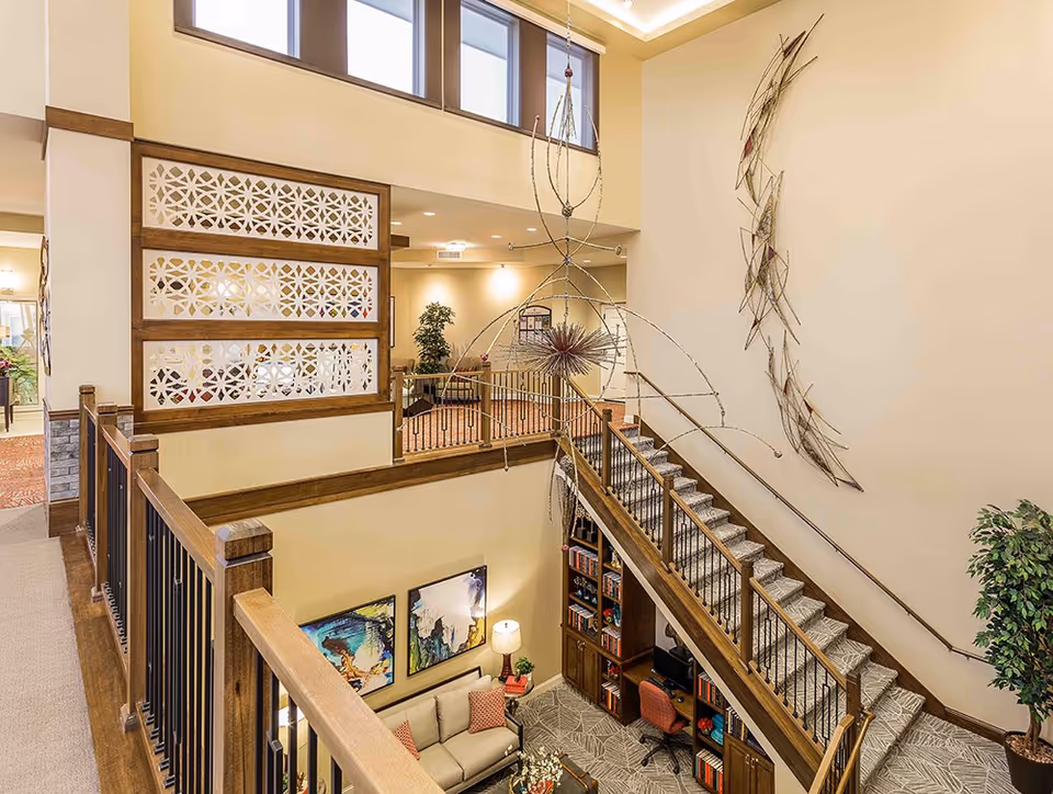 Two-story interior atrium of an assisted living facility showing a staircase, hanging metal sculpture, seating area and decorative wall art.