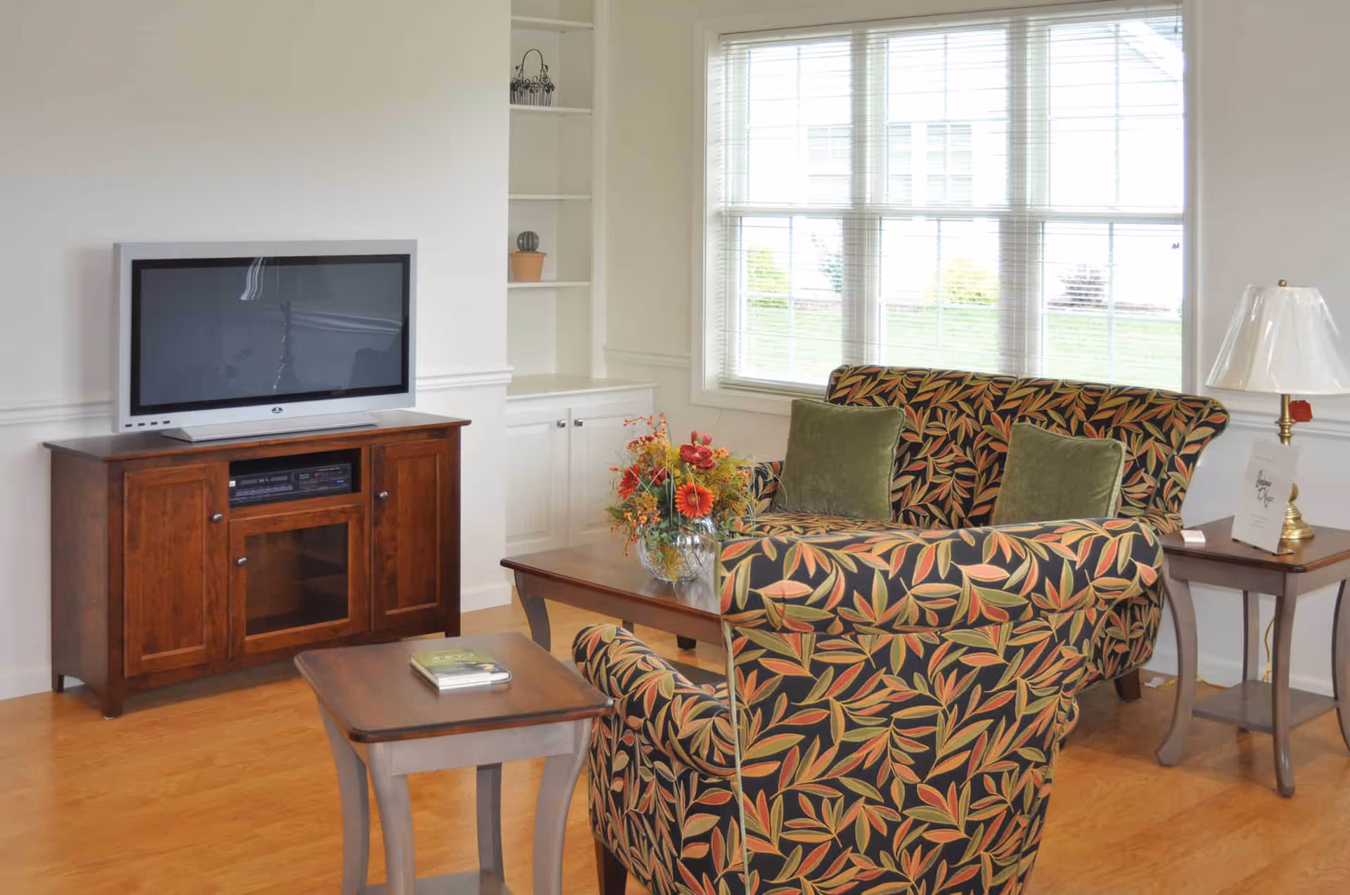 A bright living room with patterned upholstered chairs, a wooden TV cabinet, coffee table and a large window.