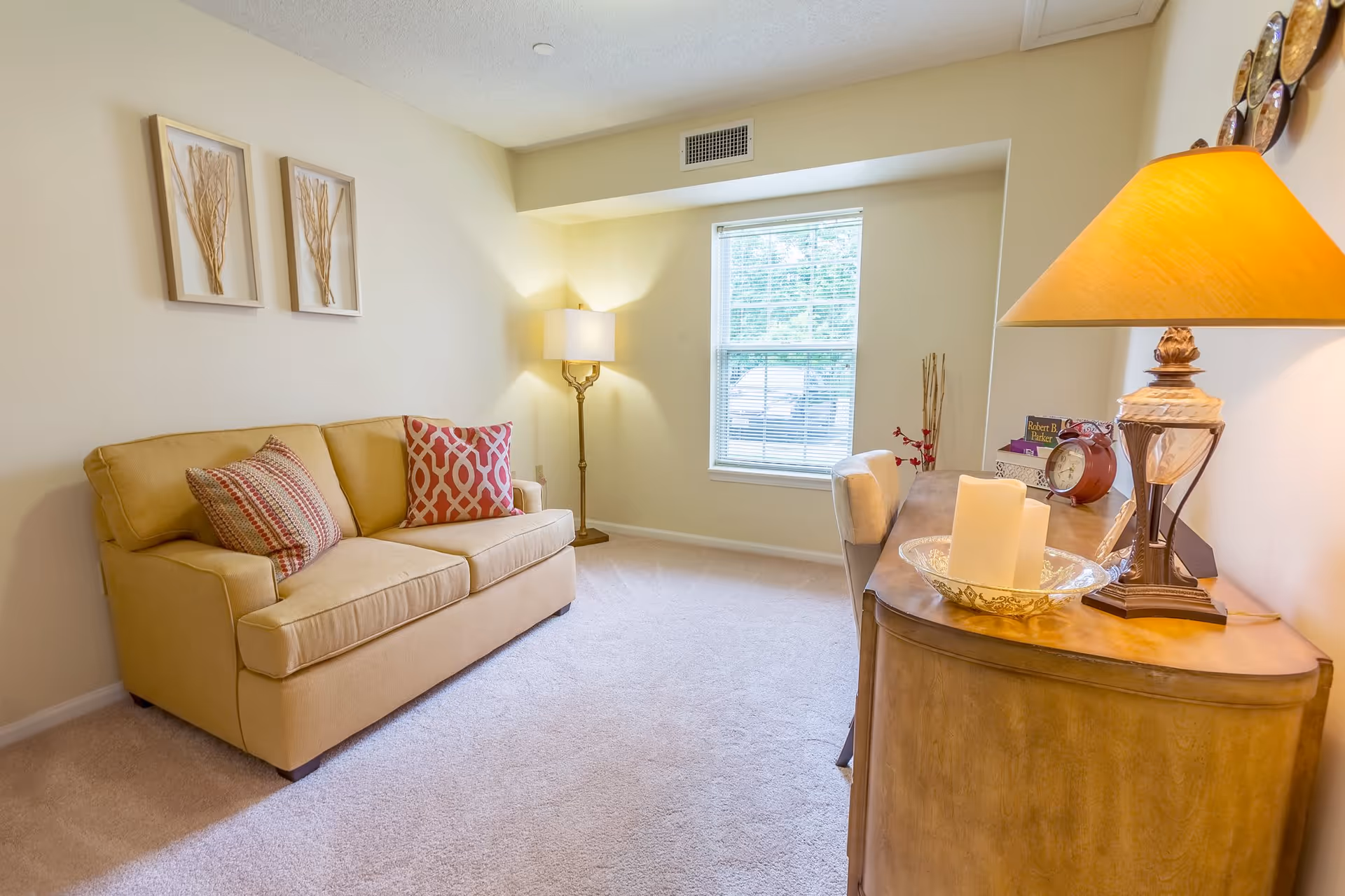 A cozy living room with a beige sofa adorned with two patterned pillows, a floor lamp in the corner, a window with blinds letting in natural light, and a wooden sideboard with a table lamp, candles, a clock, and decorative items.