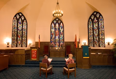 Two people seated facing the altar in a chapel with three large stained-glass windows and wooden furnishings.