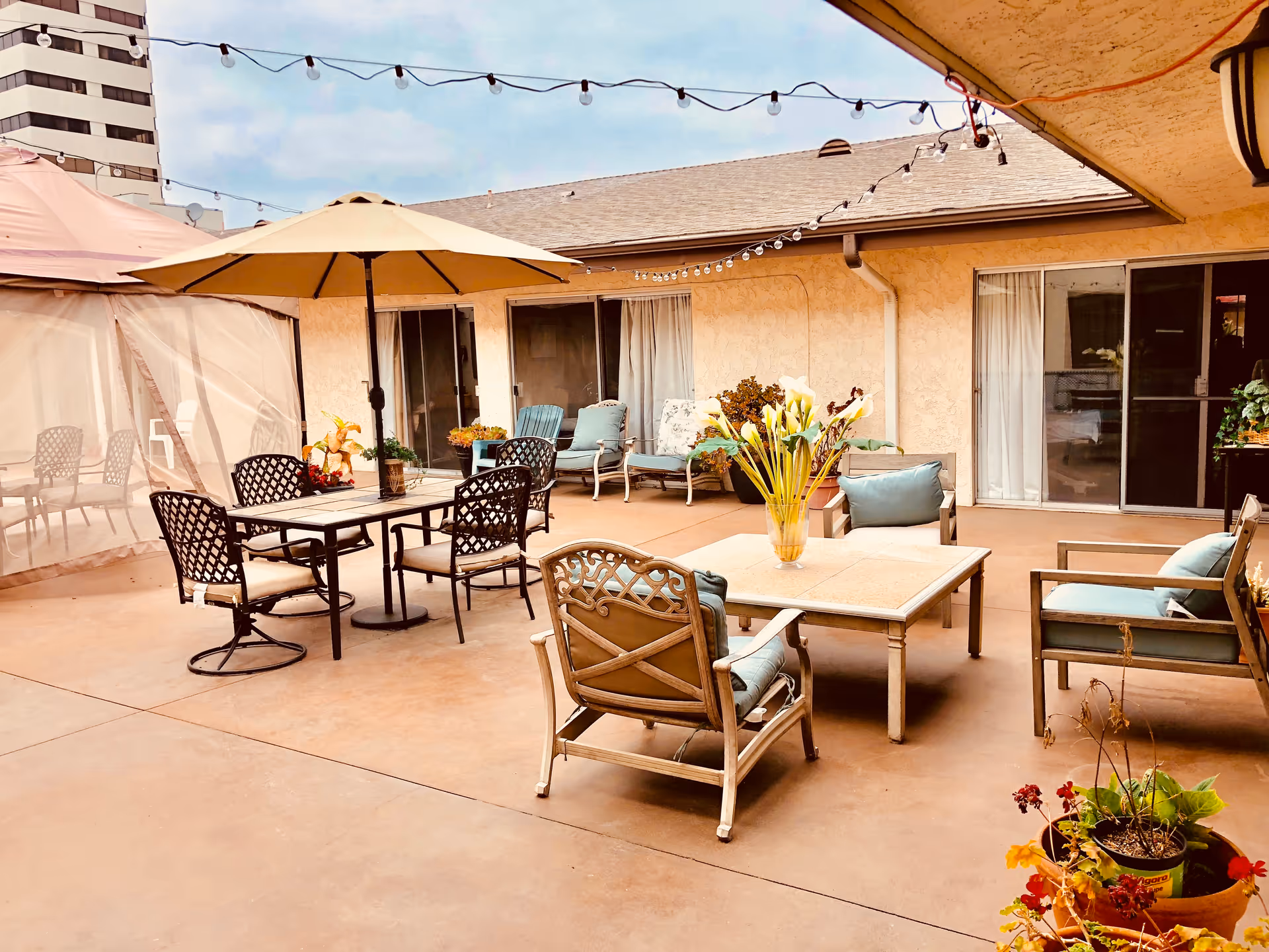 Outdoor patio area at Sea Cliff Assisted Living featuring multiple seating arrangements including cushioned chairs around a square table with a vase of flowers, a dining table with an umbrella, string lights overhead, potted plants, and sliding glass doors leading inside.