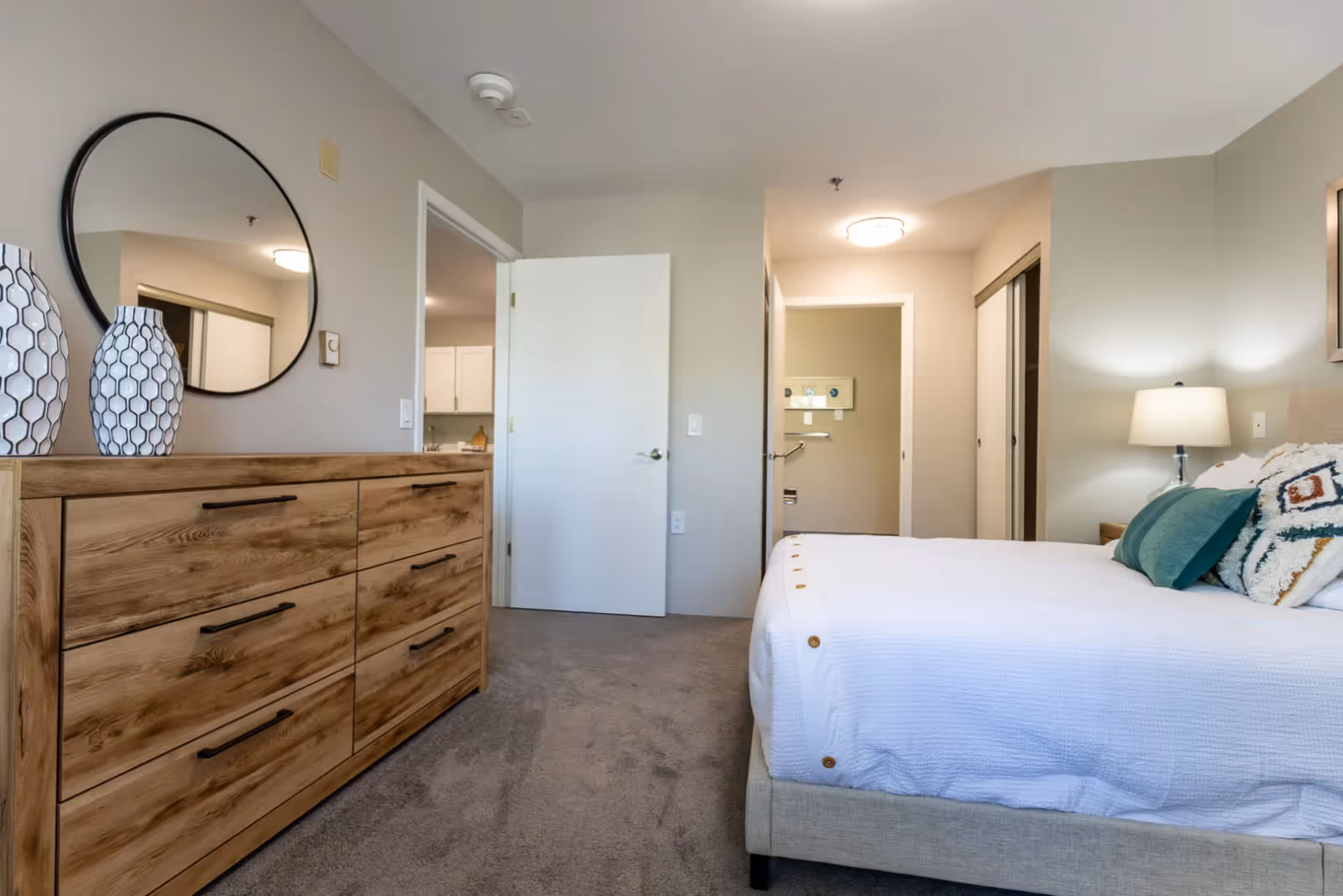 A bedroom with a beige upholstered bed featuring white bedding and decorative pillows. To the left is a wooden dresser with six drawers and two white vases with black geometric patterns on top. Above the dresser is a large round mirror. The room has beige walls and carpeted floor. There is a door open leading to another room and a hallway with a closet and a lamp on a nightstand beside the bed.