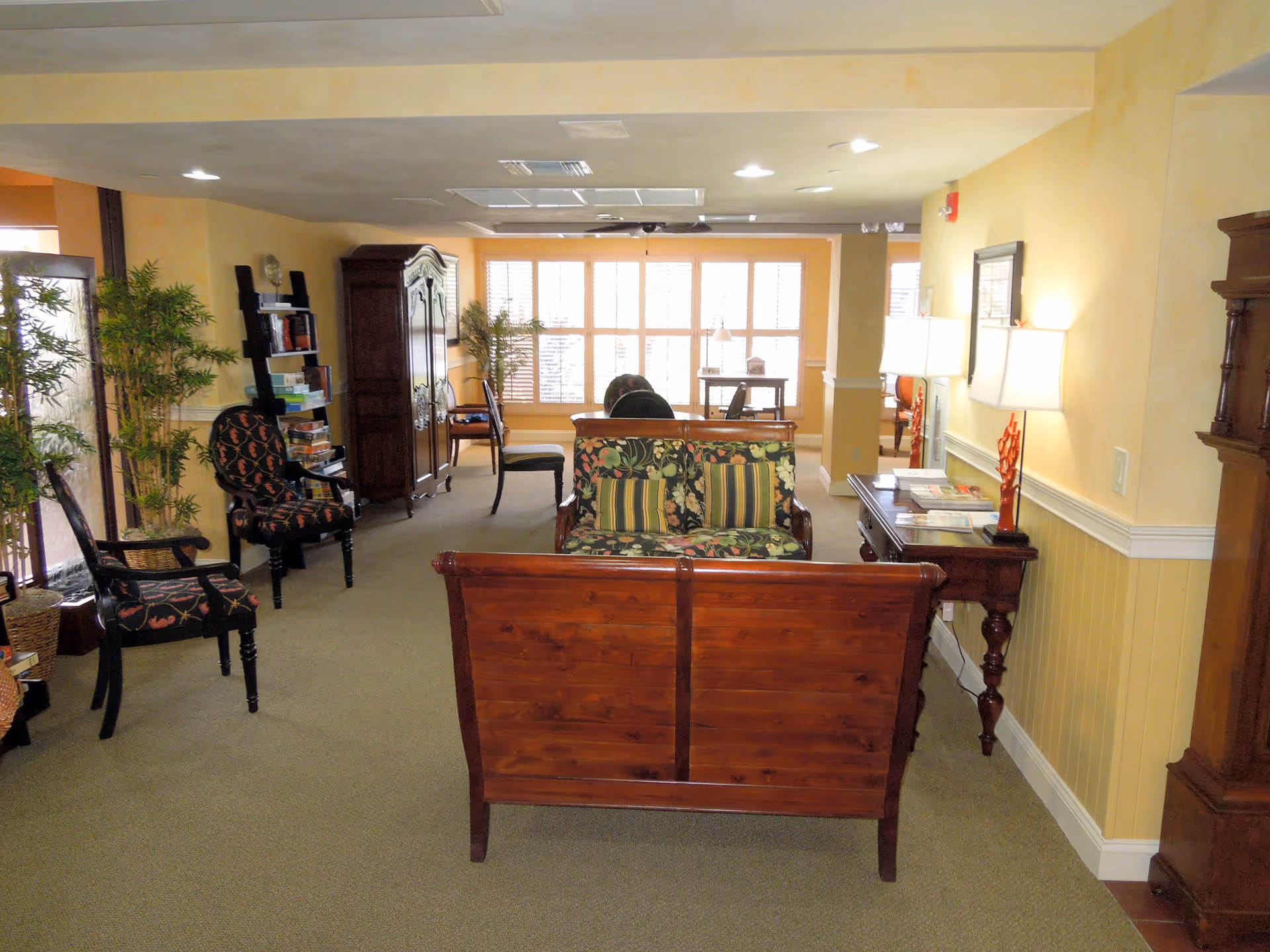 A cozy living room area with wooden furniture including a floral-patterned loveseat, chairs with dark upholstery, a wooden armoire, and a side table with lamps. The room has beige walls, carpeted floor, and large windows with white blinds letting in natural light. There are also some plants and bookshelves visible.