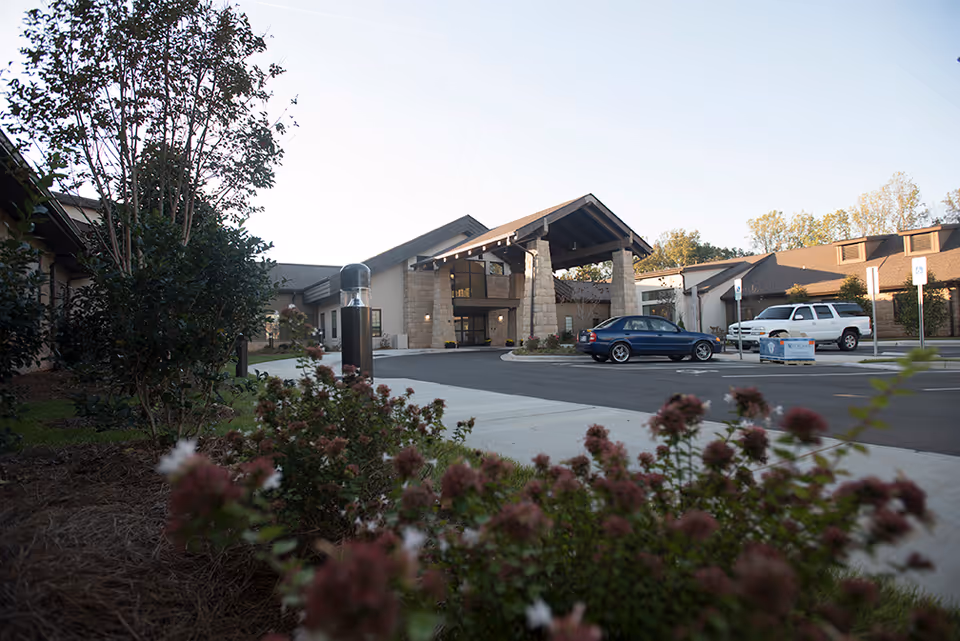 Front entrance of a single-story rehabilitation facility with a covered porte-cochere, parked cars, and landscaped foreground.