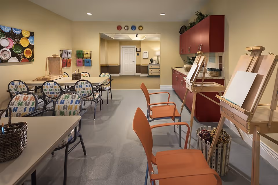 A bright and organized art activity room with several tables and chairs. There are easels with blank canvases set up along one side, colorful patterned chairs around tables, and red cabinets with plants and baskets on the countertop. The walls are painted a soft yellow, and there is a colorful painting of paint pots on one wall. The room opens into a hallway with a white door and additional furniture visible.