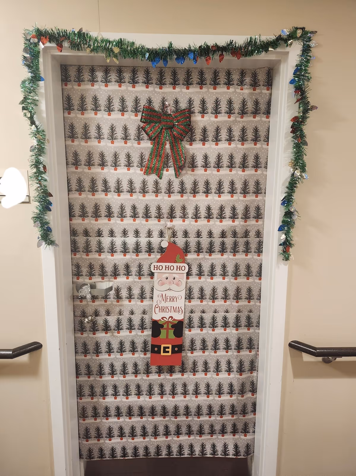 A hallway door decorated for Christmas with tree-patterned wrapping paper, garland, a large bow, and a Santa "Merry Christmas" sign.
