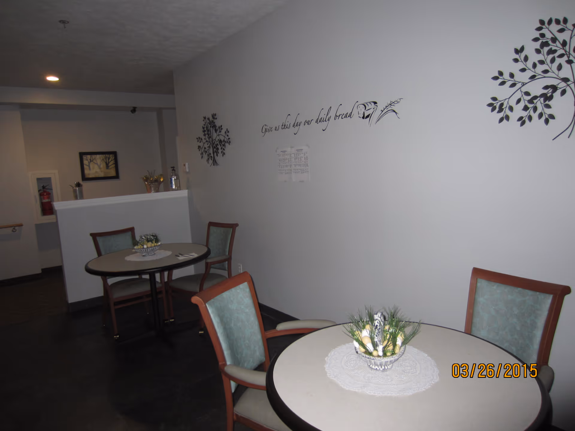 Small dining area with two round tables, each surrounded by chairs with light green upholstery. One table has a decorative centerpiece with greenery and eggs on a white doily. The wall has decorative tree decals and a quote that reads 'Give us this day our daily bread' along with an image of bread and wheat. A fire extinguisher and framed artwork are visible in the background.
