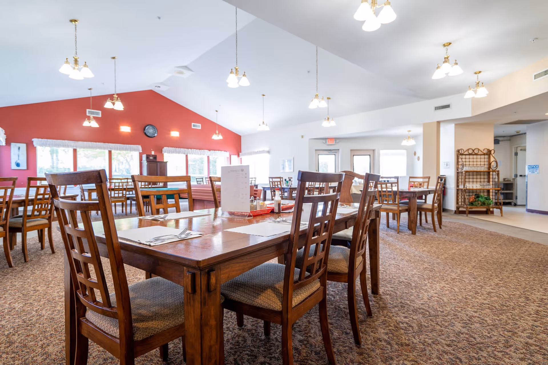 A spacious dining room with multiple wooden tables and chairs arranged neatly. The room has a high ceiling with several hanging light fixtures, a red accent wall with windows covered by white curtains, and a clock mounted on the wall. The carpeted floor and bright natural light create a welcoming atmosphere.