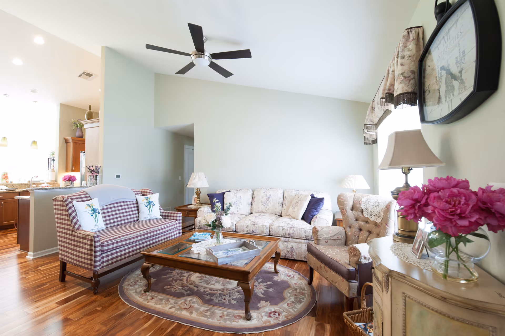 A bright and cozy living room with a floral patterned sofa, a checkered loveseat with floral pillows, a tufted armchair, and a wooden coffee table on a round rug. There are two table lamps, a ceiling fan, a wall clock, and a vase with pink flowers on a side table. The room has wooden flooring and light-colored walls, with a view into a kitchen area in the background.