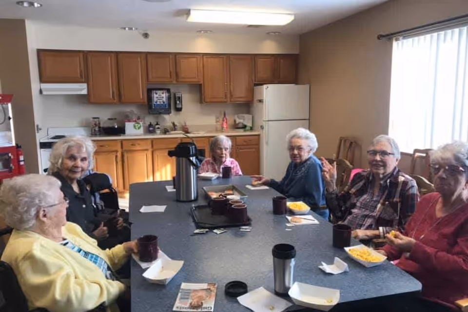 Six elderly women sitting around a rectangular table in a kitchen area, enjoying snacks and drinks. The kitchen has wooden cabinets, a white refrigerator, and a stove. There is a large window with vertical blinds letting in natural light.