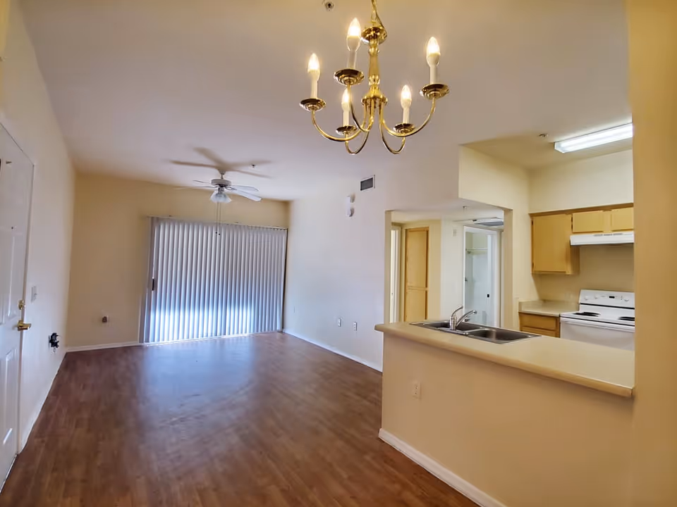 Empty interior space with wooden flooring, a ceiling fan, and vertical blinds covering a sliding glass door. A brass chandelier hangs from the ceiling above a counter with a double sink, which separates the living area from a kitchen with light wood cabinets and a white stove.