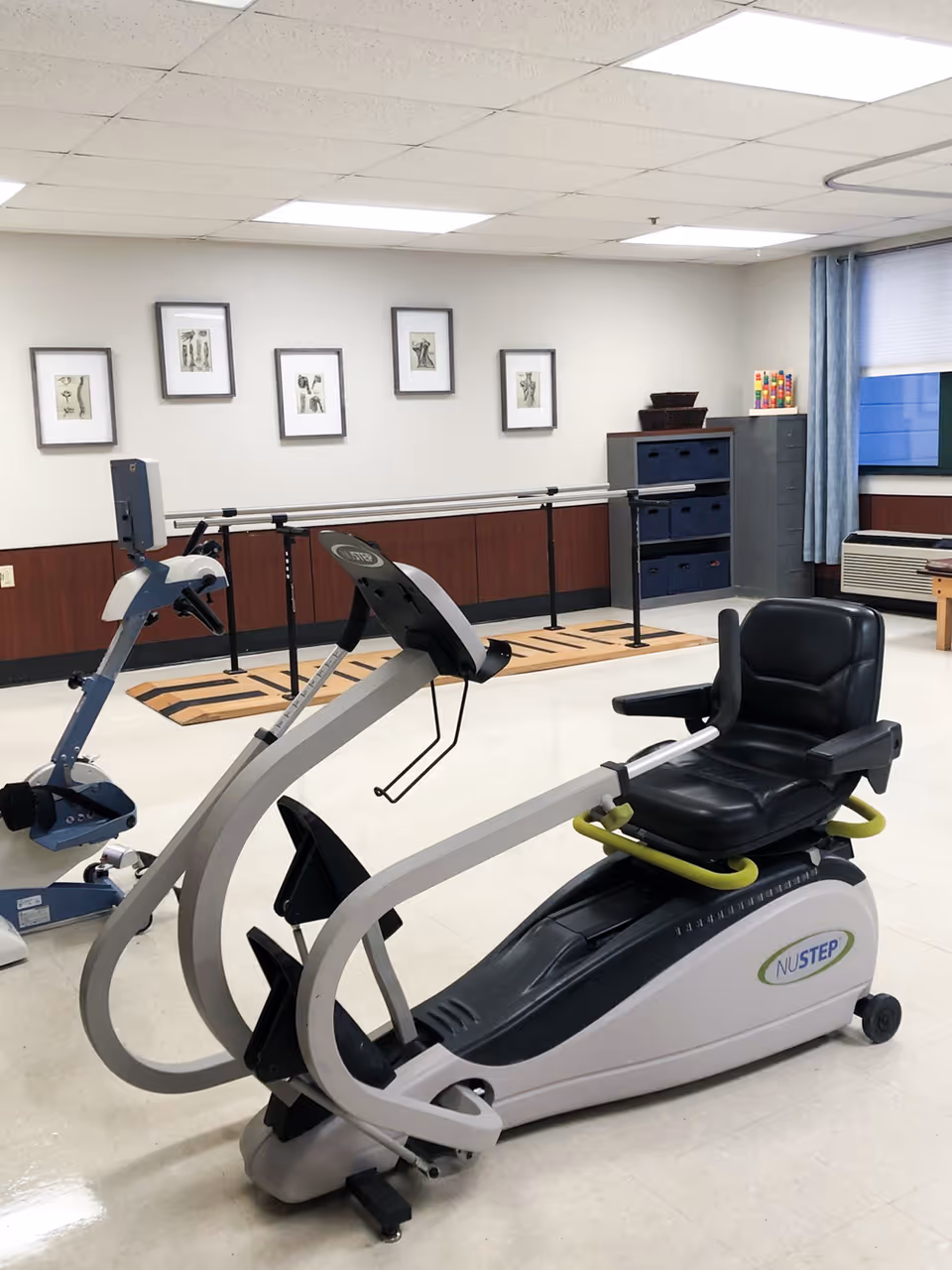 A rehabilitation or exercise room with a NuStep recumbent cross trainer and another piece of exercise equipment. The room has a wooden handrail along the wall, framed artwork, a window with blue curtains, and storage cabinets with bins.