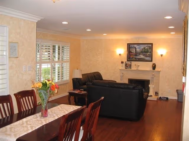 A cozy living and dining area with wooden flooring. The dining table has a vase with colorful flowers and is surrounded by wooden chairs. The living area features black leather sofas facing a white fireplace with a framed painting above it. The walls are light beige with two wall sconces providing warm lighting. A window with white shutters is visible on the left side.
