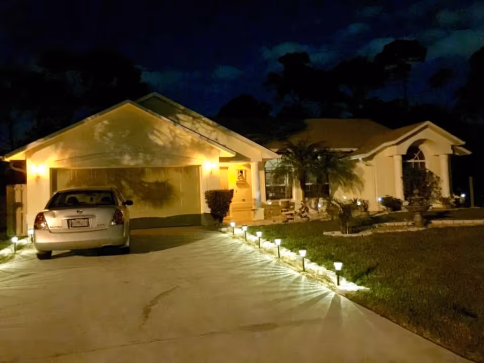 Front exterior of a single-story senior living facility at night with illuminated driveway and walkway lights and a parked car.