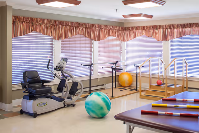 A bright therapy room with large windows covered by blinds and valances. The room contains exercise equipment including a recumbent bike, parallel bars for walking support, a set of stairs with handrails, colorful therapy balls, and padded tables with colored rods on top.