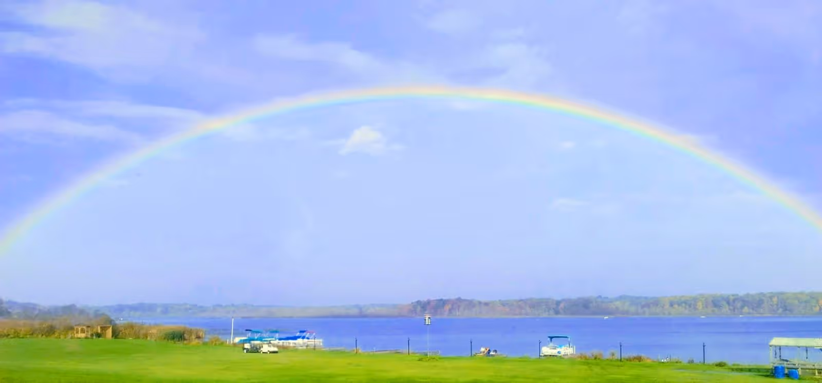 A vibrant rainbow arches across a clear blue sky over a calm lake with green grassy shore in the foreground. Several boats are docked along the lake's edge, and trees line the horizon in the distance.