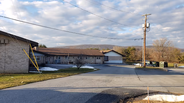 Exterior view of a single-story brick senior living building with a driveway and parking area, utility pole in front and wooded hills under a partly cloudy sky.