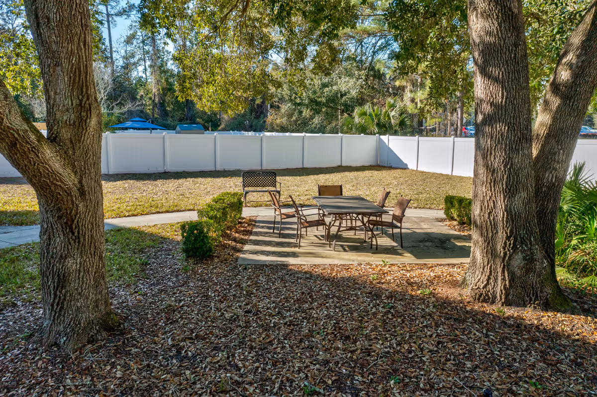 Outdoor patio area with a table and chairs set on a concrete pad beneath large trees and a white privacy fence.